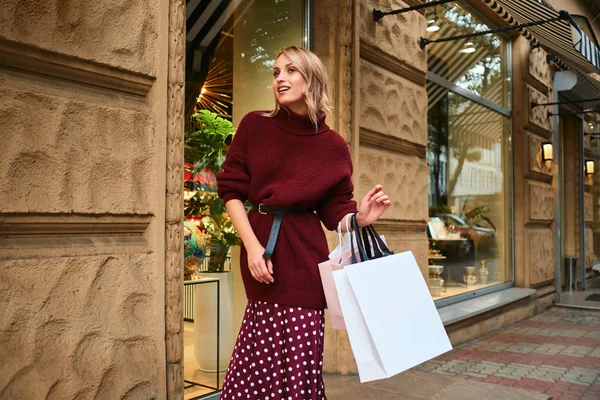 Chica rubia bastante elegante con bolsas de compras caminando soñadoramente por la calle de la ciudad — Foto de Stock