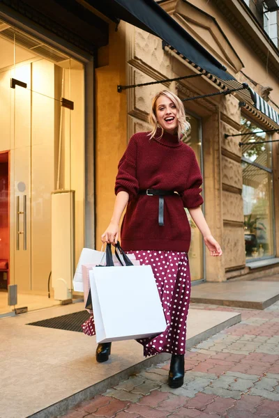 Atractiva chica rubia alegre en suéter de punto con bolsas de compras mirando felizmente en la cámara después de comprar al aire libre — Foto de Stock