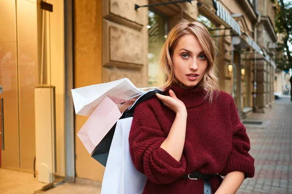 Vonzó, elegáns szőke lány kötött pulóverben, bevásárlószatyrokkal magabiztosan néz a kamerába vásárlás után a város utcájában — Stock Fotó