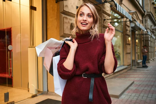 Chica rubia bastante elegante en suéter de punto con bolsas de compras mirando alegremente en la calle de la ciudad — Foto de Stock