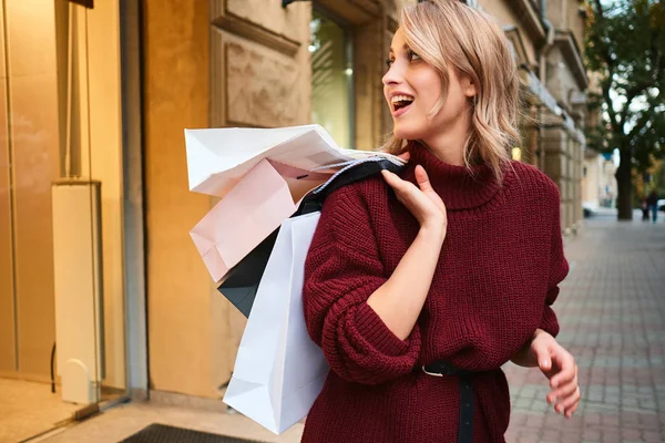 Attraktives, lässiges blondes Mädchen im Strickpullover mit Einkaufstaschen auf der Schulter, glücklich wegschauend auf der Straße der Stadt — Stockfoto