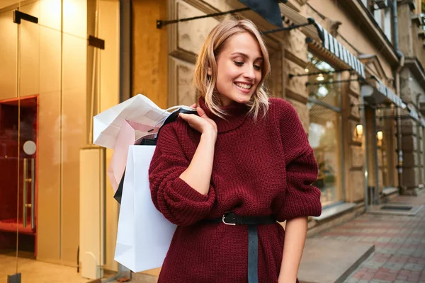 Vonzó boldog szőke lány kötött pulóverben bevásárló táskákkal vásárlás után a város utcáján — Stock Fotó