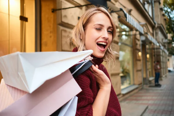 Porträt eines fröhlichen blonden Mädchens mit Einkaufstüten, das fröhlich auf der Straße herumblinzelt — Stockfoto