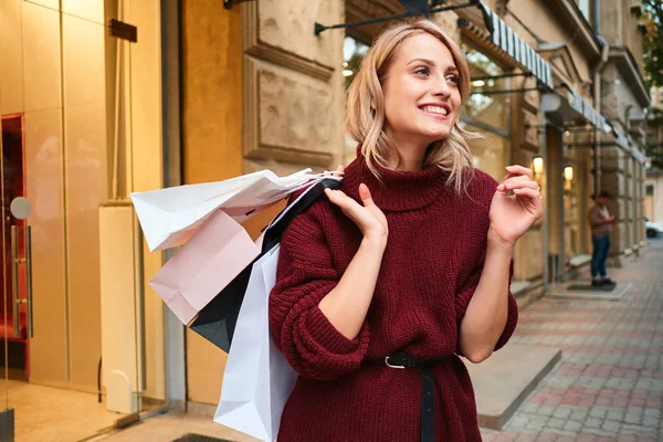 Chica rubia alegre en suéter de punto con bolsas de compras en el hombro mirando felizmente a la calle de la ciudad — Foto de Stock