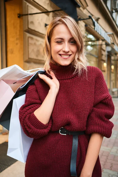 Hübsches, lächelndes blondes Mädchen mit Einkaufstüten, das nach dem Einkaufen fröhlich in die Kamera zwinkert — Stockfoto