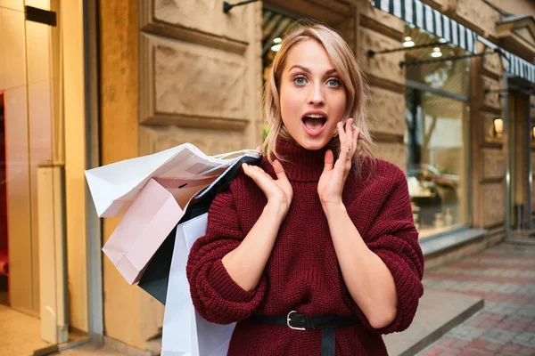 Porträt eines überraschten, stylischen blonden Mädchens mit Einkaufstüten, das staunend in die Kamera auf der Straße der Stadt blickt — Stockfoto