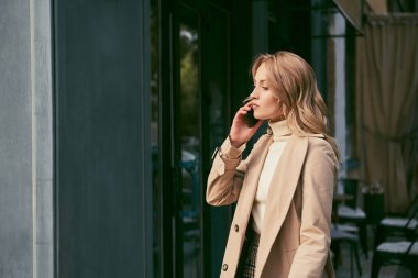 Trençkotlu çekici sarışının yan görüntüsü açık havada cep telefonuyla düşünceli bir şekilde konuşuyor.