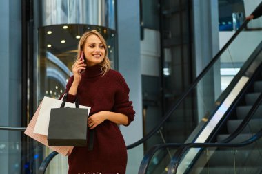 Örgü kazaklı neşeli sarışın kız alışveriş merkezinde cep telefonuyla konuşuyor.