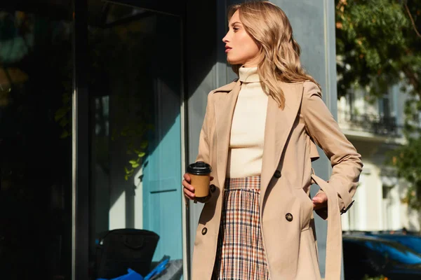 Side view of attractive stylish blond girl in beige coat with coffee to go walking on city street — Zdjęcie stockowe