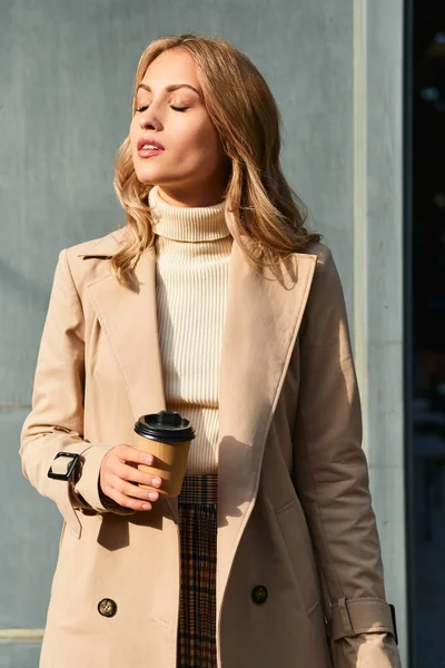 Mooi zonnig blond meisje in beige jas met koffie te gaan dromerig staan buiten — Stockfoto