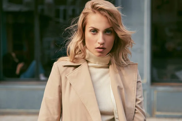 Beautiful serious blond girl in trench coat confidently looking in camera on street — Stock Photo, Image