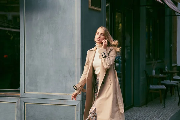 Beautiful blond girl in stylish trench coat talking on cellphone on street — Stock Photo, Image