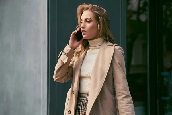 Beautiful casual blond girl in trench coat talking on cellphone thoughtfully looking away outdoor — Stock Photo, Image