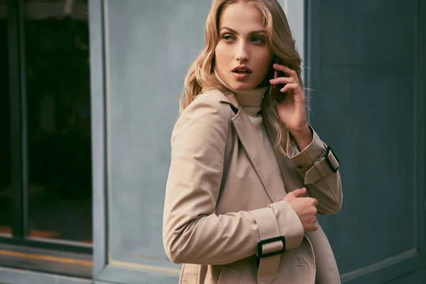 Attractive casual blond girl in trench coat talking on cellphone sensually looking away outdoor — Zdjęcie stockowe