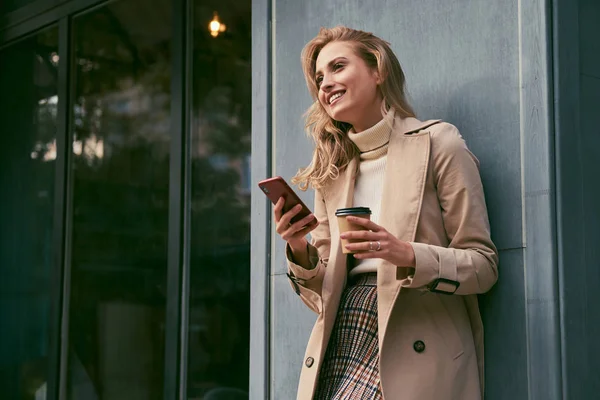 Atraktivní veselá blondýnka v trenč kabát s kávou jít šťastně pomocí mobilu venkovní — Stock fotografie
