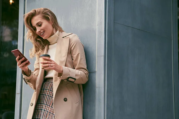 Pěkně usměvavá blond dívka ve stylové trenč kabát s kávou jít radostně pomocí mobilního telefonu venku — Stock fotografie