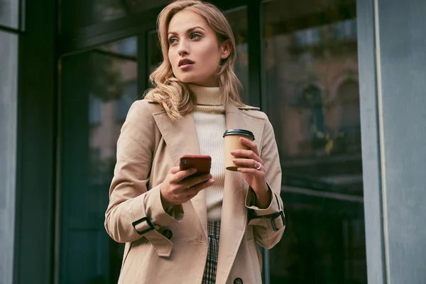 Aantrekkelijk casual blond meisje in stijlvolle trench jas met koffie — Stockfoto