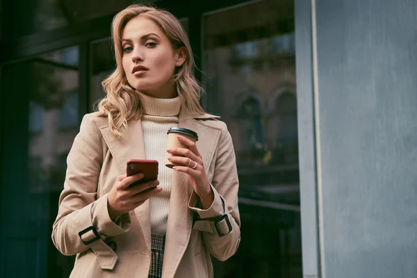 Piękny casual blondynka dziewczyna w płaszcz z kawą iść i telefon pewnie patrząc w aparat na zewnątrz — Zdjęcie stockowe