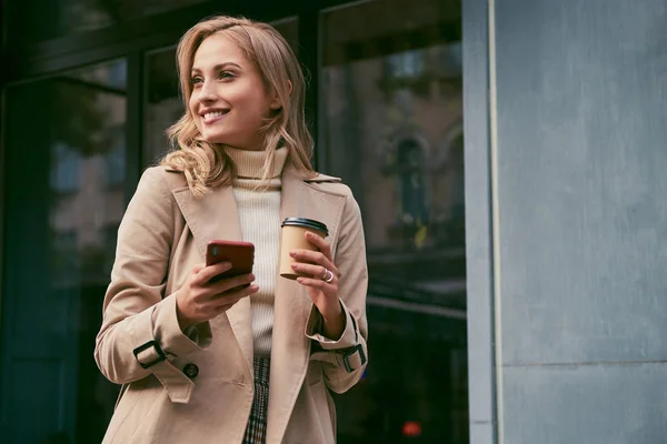 Menarik tersenyum santai gadis pirang dalam mantel parit dengan kopi untuk pergi dan ponsel gembira melihat keluar rumah — Stok Foto