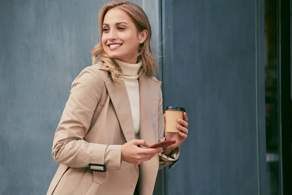 Довольно улыбается случайная блондинка в плаще с кофе идти и сотовый телефон счастливо глядя в сторону на открытом воздухе — стоковое фото