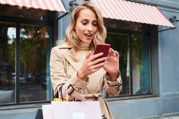 Attractive surprised blond girl in stylish trench coat with shopping bags happily using cellphone on street — Stock Photo, Image
