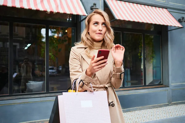 Attraktive kokette blonde Mädchen in stylischem Trenchcoat mit Einkaufstüten verträumt mit Handy auf der Straße — Stockfoto