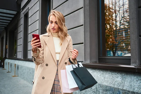 Menina loira elegante bonita em casaco bege com sacos de compras usando celular na rua — Fotografia de Stock