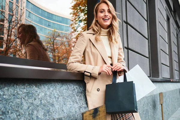 Menina loira elegante muito alegre no casaco bege com sacos de compras olhando alegremente na câmera na rua — Fotografia de Stock