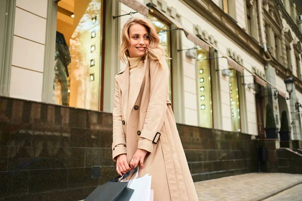Csinos mosolygós, elegáns, szőke lány bézs kabátban, bevásárlószatyrokkal, örömmel néz félre a város utcáján. — Stock Fotó