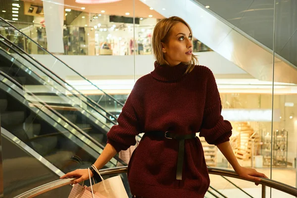 Piękny stylowy blondynka dziewczyna w swetrze z torby na zakupy Drethe stojąc w nowoczesnym centrum handlowym — Zdjęcie stockowe