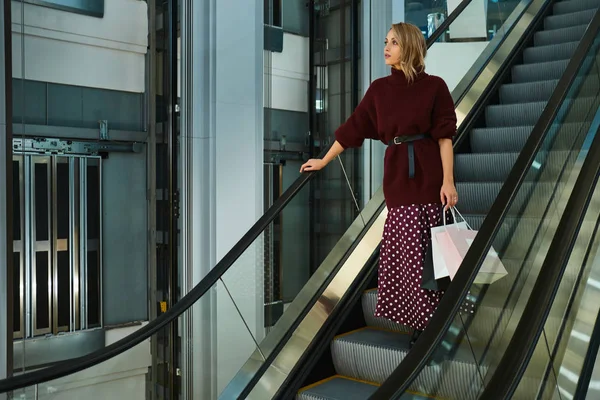 Bella ragazza bionda elegante con borse della spesa pensieroso guardando lontano sulla scala mobile nel moderno centro commerciale — Foto Stock