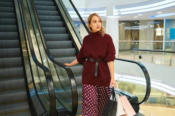Belle fille blonde avec des sacs à provisions sur l'escalator en regardant attentivement loin dans le centre commercial moderne — Photo