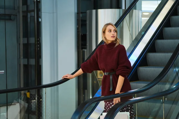 Hermosa chica rubia con bolsas de compras en escaleras mecánicas mirando hacia otro lado en el moderno centro comercial — Foto de Stock