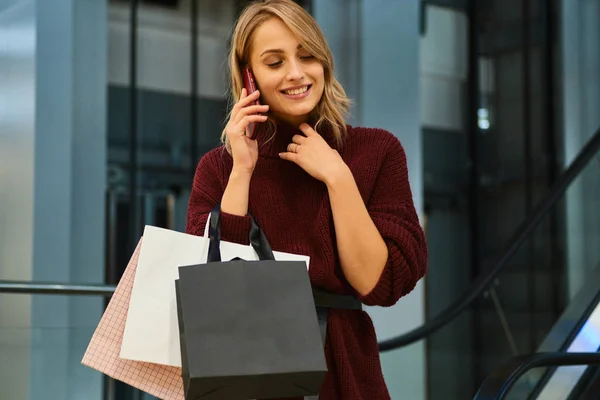 Hübsches, lächelndes blondes Mädchen im Strickpullover, das in einem Einkaufszentrum fröhlich mit dem Handy spricht — Stockfoto