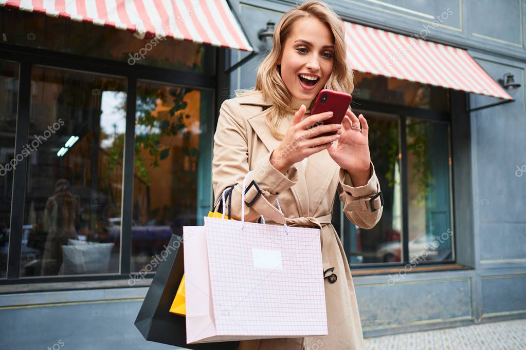 Attractive excited blond girl in stylish trench coat with shopping bags joyfully using cellphone on city street