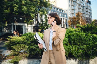Bej takım elbiseli, dizüstü bilgisayarlı, şehirdeki caddede cep telefonuyla konuşan genç ve güzel bir iş kadını.
