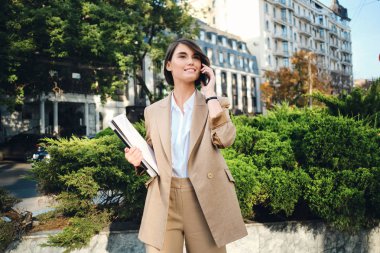 Bej takım elbiseli, dizüstü bilgisayarlı, cep telefonuyla konuşan güzel bir iş kadını.