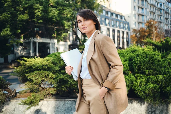 Giovane bella donna d'affari in abito beige in piedi con computer portatile intensamente guardando in macchina fotografica sulla strada della città — Foto Stock