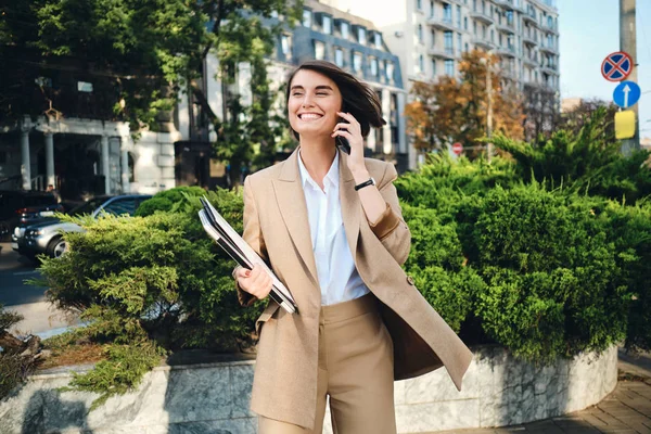 Giovane donna d'affari allegra in abito beige con laptop che parla felicemente sul cellulare sulla strada della città — Foto Stock