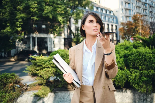 Giovane donna d'affari attraente in abito beige con computer portatile che registra con attenzione il messaggio vocale sul cellulare sulla strada della città — Foto Stock