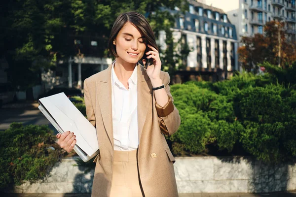 Giovane bella donna d'affari in abito beige con computer portatile felicemente parlando sul cellulare sulla strada della città — Foto Stock