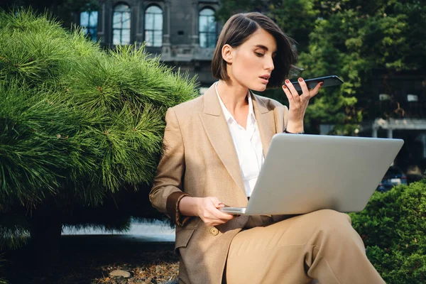 Молодая красивая стильная деловая женщина работает на ноутбуке записи голосовых сообщений на открытом воздухе — стоковое фото