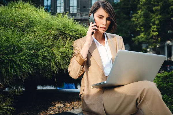 Молодая серьезная стильная деловая женщина разговаривает по мобильному телефону и работает на открытом воздухе ноутбука — стоковое фото