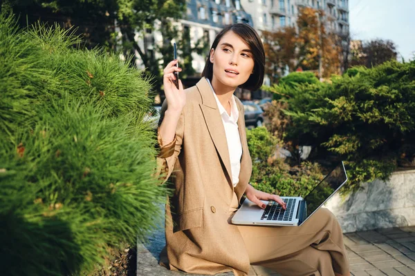 年轻迷人的时髦女商人，身穿米色西服，带着手机在户外笔记本电脑上工作 — 图库照片