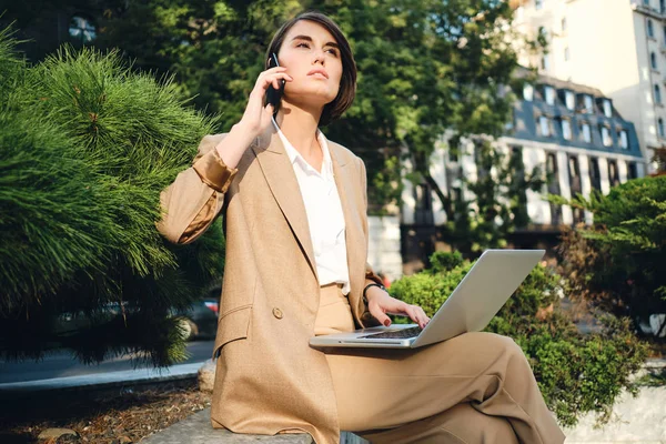 Молодая вдумчивая деловая женщина разговаривает по мобильному телефону, работая с ноутбуком на городской улице — стоковое фото