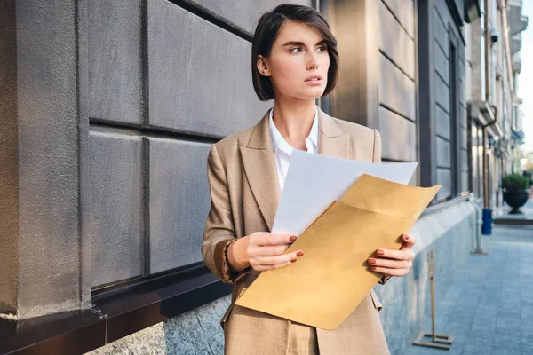 Giovane donna d'affari seria in giacca e cravatta intensamente guardando lontano lavorando con i documenti sulla strada — Foto Stock