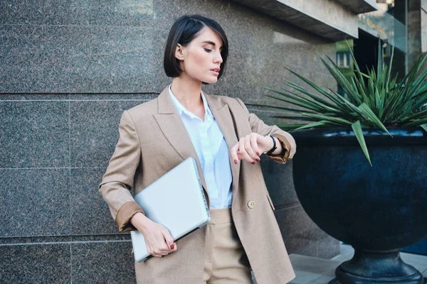 Młoda, pewna siebie stylowa bizneswoman w beżowym garniturze z laptopem oglądająca czas na zegarku outdoor — Zdjęcie stockowe