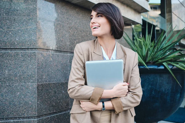 Giovane donna d'affari elegante piuttosto sorridente con il computer portatile gioiosamente guardando da parte sulla strada — Foto Stock