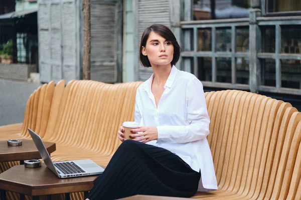 Jovem mulher de negócios elegante com laptop e café cuidadosamente trabalhando no café na rua — Fotografia de Stock