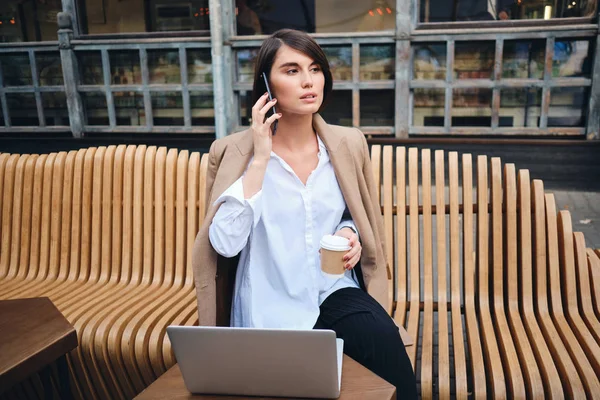 Молодая уверенная в себе стильная деловая женщина с ноутбуком разговаривает по телефону во время кофе-брейка в кафе на улице — стоковое фото